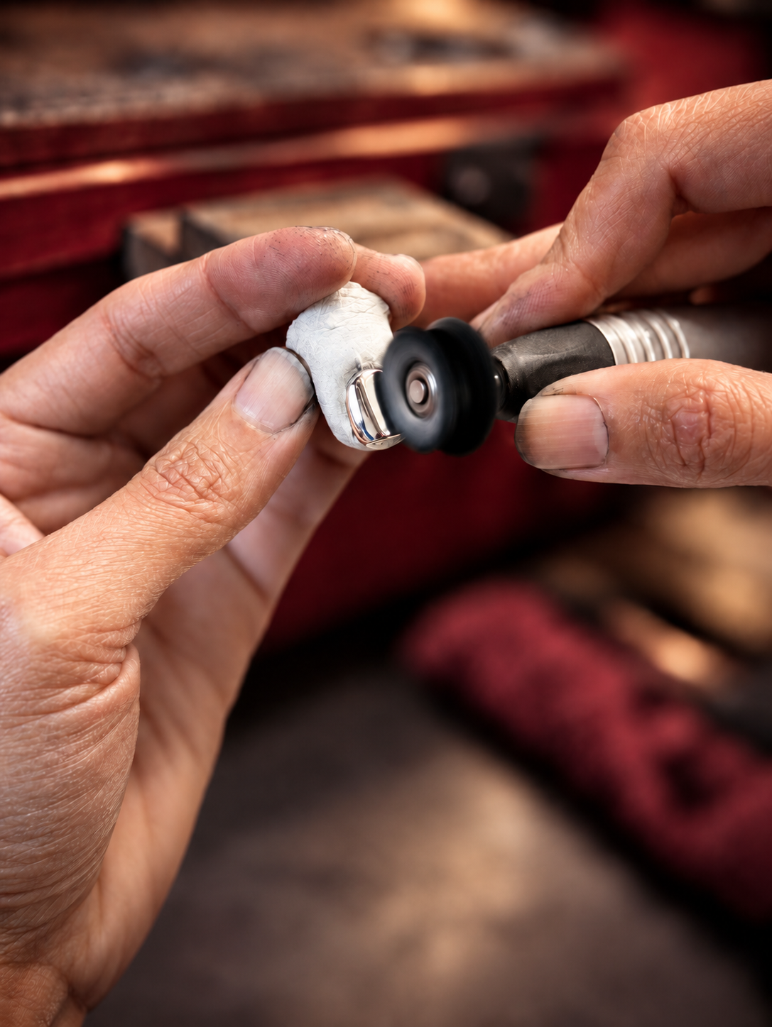 Close-up of a jeweler's hands using a rotary diamond grinding wheel to polish a small silver ring on a white stone, with a blurred workshop background.
