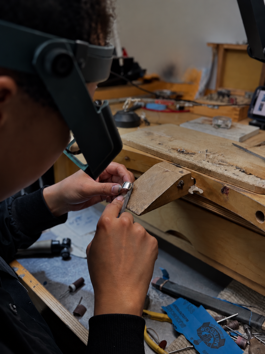 A close-up lifestyle shot of a jeweler wearing a magnifying loupe and working at a wooden jeweler's bench, holding a small metal ring under magnification.