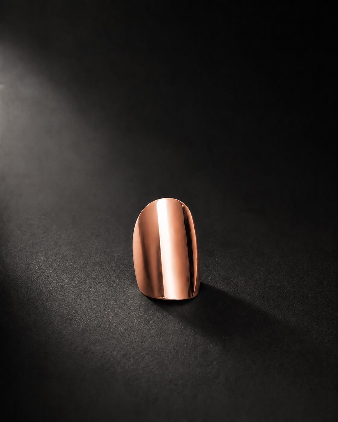 A single metallic rose 14k gold nail tip or press-on nail standing upright on a dark textured surface with dramatic spotlighting.