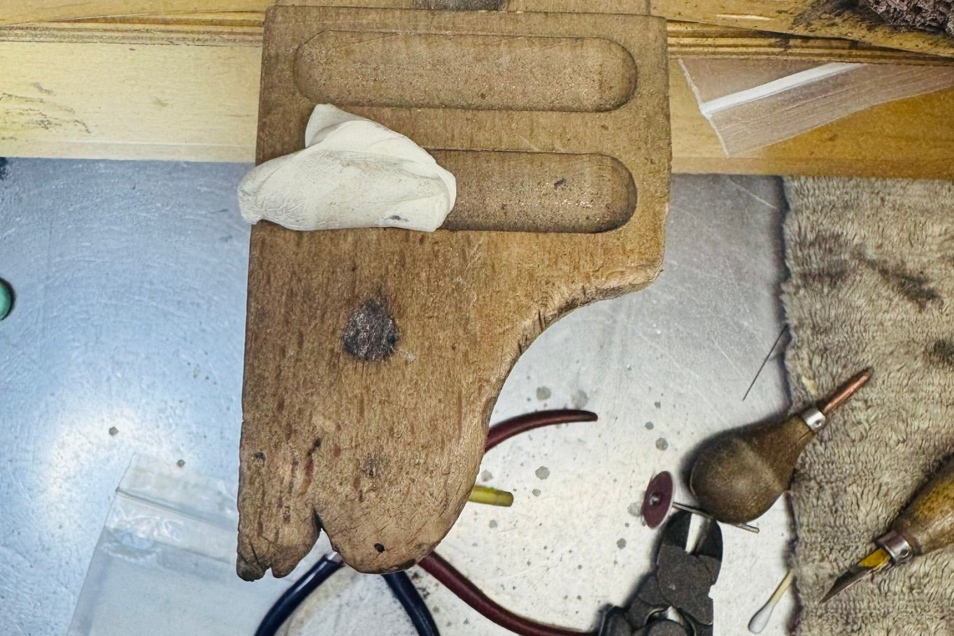 A close-up of a wooden jewelry organizer or tool holder mounted on a workbench, holding a white cloth, with pliers, a burlap-wrapped tool, and other jewelry-making tools scattered around on a metal work surface.